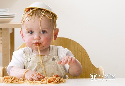 Messy baby boy in high chair with bowl of spaghetti on head